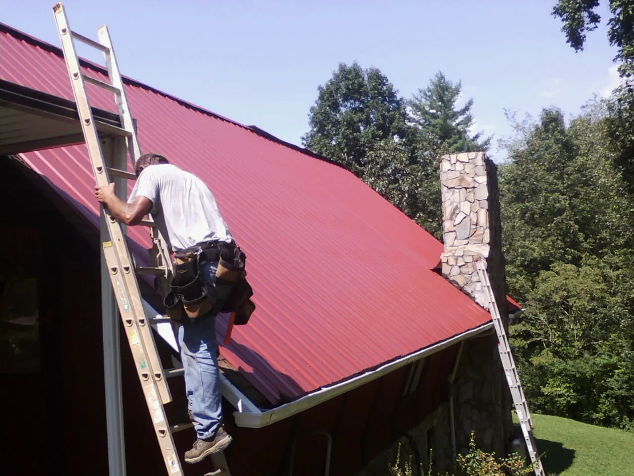 Roofer on a ladder beside a red metal roof and stone chimney — Soffit & Fascia Repair by Roofing Brothers in Ardmore